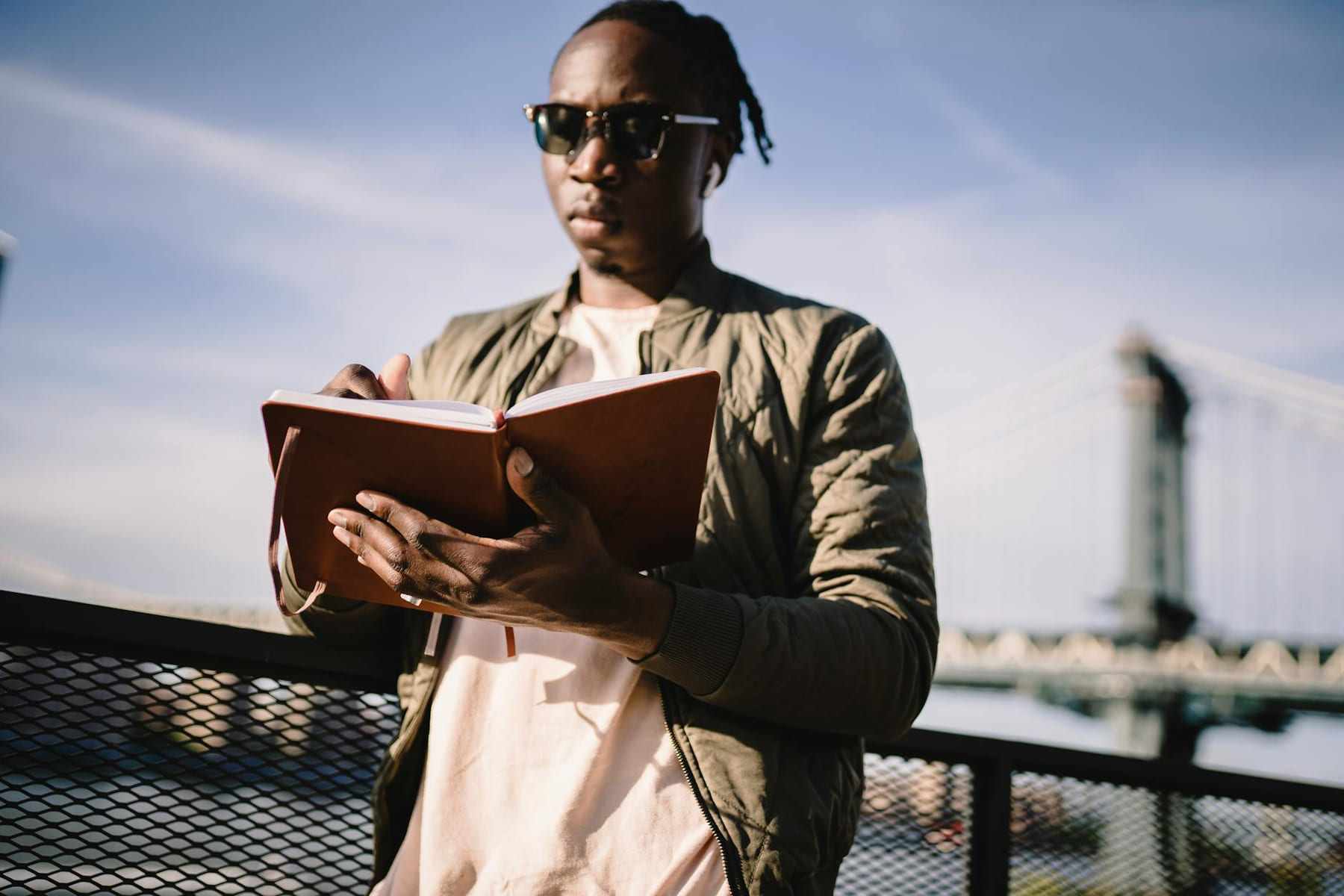 Low angle of serious African American guy in sunglasses and true wireless earphones writing in notebook while standing near fence on bridge over city river and listening to music on Inner Progress Project